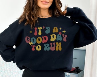 May include: A navy blue sweatshirt with the text "IT'S A GOOD DAY TO RUN" in retro-style, multi-colored lettering. The design includes stars and flowers. The sweatshirt is a casual, comfortable garment.