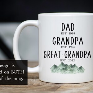 K&ouml;nnte beinhalten: Wei&szlig;e Keramiktasse mit schwarzem Text, der "DAD EST. 1988 GRANDPA EST. 1996 GREAT-GRANDPA EST. 2022" lautet, mit einer gr&uuml;nen Aquarell-Bergkette am Boden.