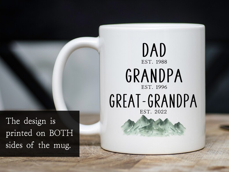 K&ouml;nnte beinhalten: Wei&szlig;e Keramiktasse mit schwarzem Text, der "DAD EST. 1988 GRANDPA EST. 1996 GREAT-GRANDPA EST. 2022" lautet, mit einer gr&uuml;nen Aquarell-Bergkette am Boden.