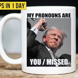 May include: White ceramic coffee mug with a black and white image of a man with a raised fist. The text "MY PRONOUNS ARE YOU/MISSED" is printed on the mug.