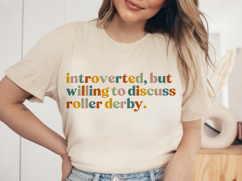 K&ouml;nnte beinhalten: Ein wei&szlig;es T-Shirt mit dem Text "introvertiert, aber bereit, &uuml;ber Roller Derby zu diskutieren." in bunten Buchstaben.
