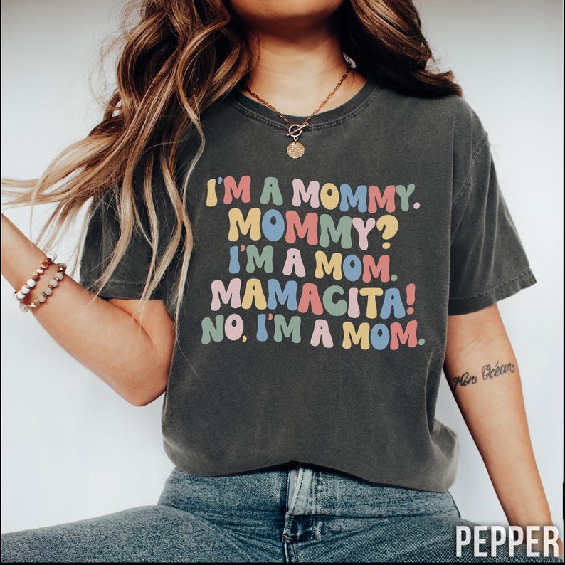 K&ouml;nnte beinhalten: Dunkelgraues T-Shirt mit dem Text "I'M A MOMMY. MOMMY? I'M A MOM. MAMACITA! NO, I'M A MOM." in bunten Buchstaben. Das Shirt ist ein l&auml;ssiger Stil, geeignet f&uuml;r den Alltag.