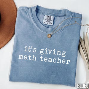 May include: A blue jean colored t-shirt with the text "it's giving math teacher" in white. A gold necklace with a pendant is draped across the shirt. A brown hat and dried plant are in the background.