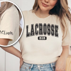 Op de afbeelding: Een cr&egrave;mekleurig T-shirt met de tekst "LACROSSE MOM" in zwarte blokletters. De mouw heeft een zwarte lacrosse-stick-afbeelding en de tekst "Liam" in zwarte schrijfletters.