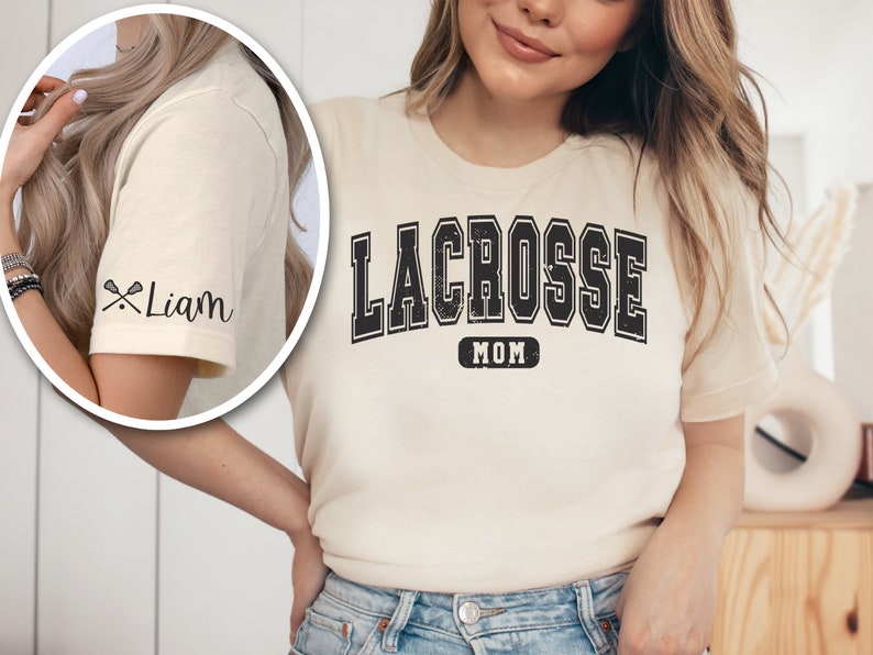 Op de afbeelding: Een cr&egrave;mekleurig T-shirt met de tekst "LACROSSE MOM" in zwarte blokletters. De mouw heeft een zwarte lacrosse-stick-afbeelding en de tekst "Liam" in zwarte schrijfletters.