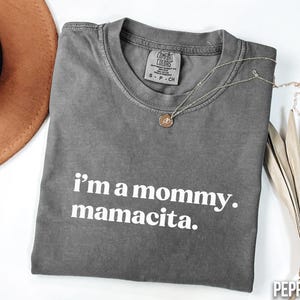 May include: A grey Comfort Colors t-shirt with the text "i'm a mommy. mamacita." in white. A gold necklace with a pendant is draped across the shirt. A brown hat is visible in the upper left corner.