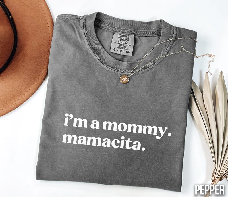 May include: A grey Comfort Colors t-shirt with the text "i'm a mommy. mamacita." in white. A gold necklace with a pendant is draped across the shirt. A brown hat is visible in the upper left corner.