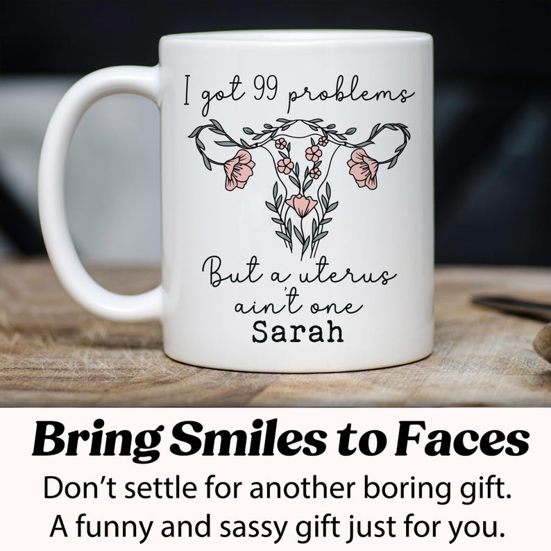 Pu&ograve; includere: Una tazza in ceramica bianca con un contorno nero di un utero con fiori rosa. La tazza recita "I got 99 problems but a uterus ain't one Sarah".