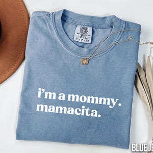 May include: A blue jean Comfort Colors t-shirt with the text "i'm a mommy. mamacita." in white. A gold necklace with a pendant is draped across the shirt. A brown hat and dried plant are in the background.