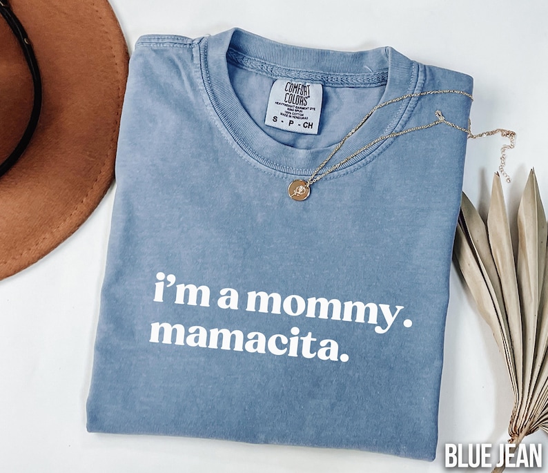 May include: A blue jean Comfort Colors t-shirt with the text "i'm a mommy. mamacita." in white. A gold necklace with a pendant is draped across the shirt. A brown hat and dried plant are in the background.