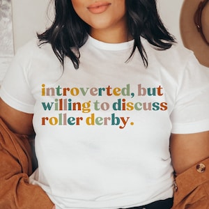 K&ouml;nnte beinhalten: Wei&szlig;es T-Shirt mit dem Text "introvertiert, aber bereit, &uuml;ber Roller Derby zu diskutieren." in bunten Buchstaben.