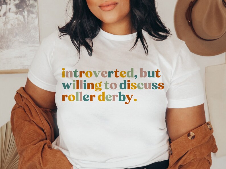 K&ouml;nnte beinhalten: Wei&szlig;es T-Shirt mit dem Text "introvertiert, aber bereit, &uuml;ber Roller Derby zu diskutieren." in bunten Buchstaben.