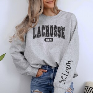 Op de afbeelding: Grijs sweatshirt met de tekst "Lacrosse Mom" op de voorkant en de tekst "Liam" met een lacrosse-stick afbeelding op de mouw.