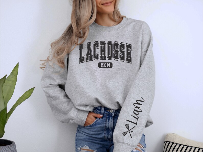 Op de afbeelding: Grijs sweatshirt met de tekst "Lacrosse Mom" op de voorkant en de tekst "Liam" met een lacrosse-stick afbeelding op de mouw.
