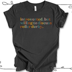 K&ouml;nnte beinhalten: Ein dunkelgraues T-Shirt mit dem Text "introvertiert, aber bereit, &uuml;ber Roller Derby zu diskutieren." in bunten Buchstaben.