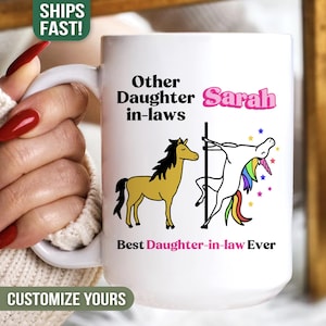 May include: White ceramic mug with the text "Other Daughter in-laws Sarah Best Daughter-in-law Ever" and illustrations of a horse and a unicorn. The mug is held by a person with red nail polish.