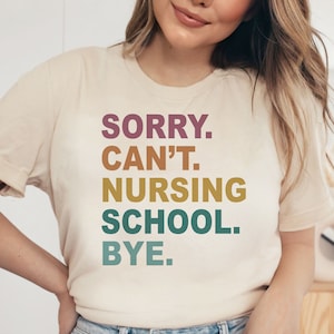 May include: Cream-colored t-shirt with the text "SORRY. CAN'T. NURSING SCHOOL. BYE." in a stacked, colorful font. The shirt has short sleeves and a crew neck. The text is in shades of purple, brown, gold, teal, and green.