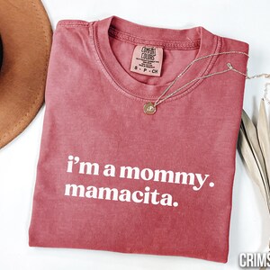 May include: Crimson t-shirt with the text "i'm a mommy. mamacita." A gold necklace with a pendant rests on the shirt. The shirt has a Comfort Colors tag. A brown hat is visible in the upper left corner.