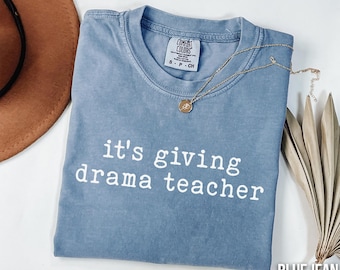 May include: A blue jean Comfort Colors t-shirt with the text "it's giving drama teacher" in white. A gold necklace with a pendant is draped across the shirt. A brown hat and dried leaves are in the background.