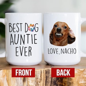 May include: White ceramic coffee mug with a black paw print and the text "Best Dog Auntie Ever" on the front. The back of the mug has a photo of a brown dachshund with the text "Love, Nacho".