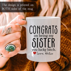K&ouml;nnte beinhalten: Wei&szlig;e Keramiktasse mit schwarzem Text, der "Congrats on being my sister you lucky bitch. Love, Katie" lautet. Ein rotes Herz befindet sich neben dem Wort "Love".