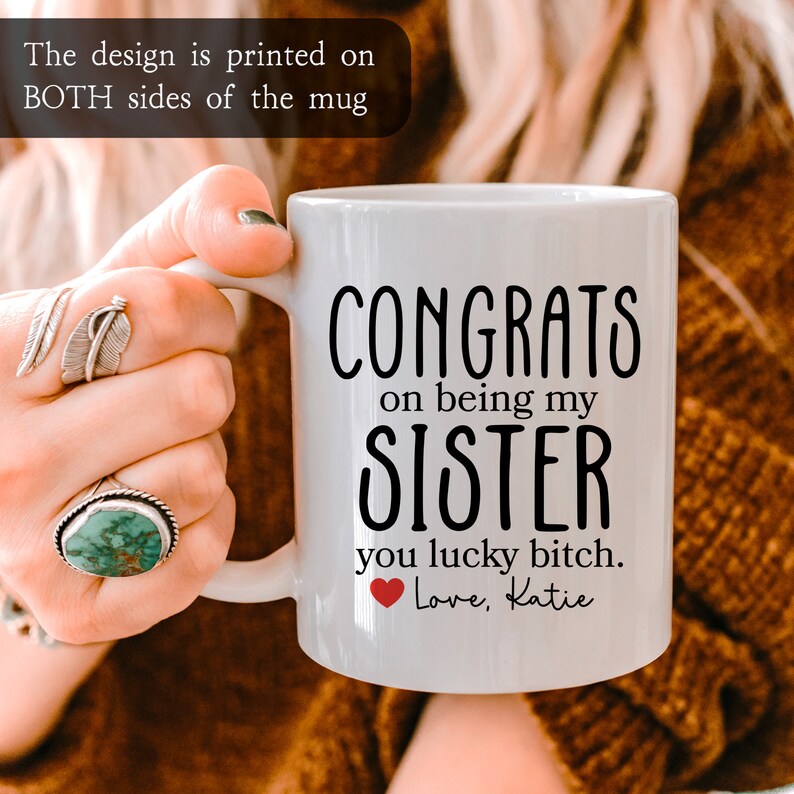 K&ouml;nnte beinhalten: Wei&szlig;e Keramiktasse mit schwarzem Text, der "Congrats on being my sister you lucky bitch. Love, Katie" lautet. Ein rotes Herz befindet sich neben dem Wort "Love".