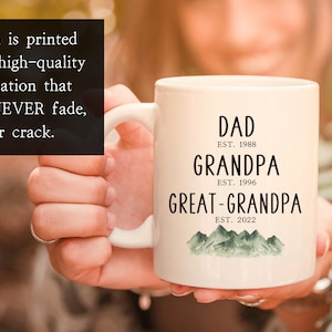 K&ouml;nnte beinhalten: Wei&szlig;e Keramiktasse mit schwarzem Text, der "DAD EST. 1988 GRANDPA EST. 1996 GREAT-GRANDPA EST. 2022" lautet, mit einem gr&uuml;nen Aquarell-Bergketten-Design am Boden.