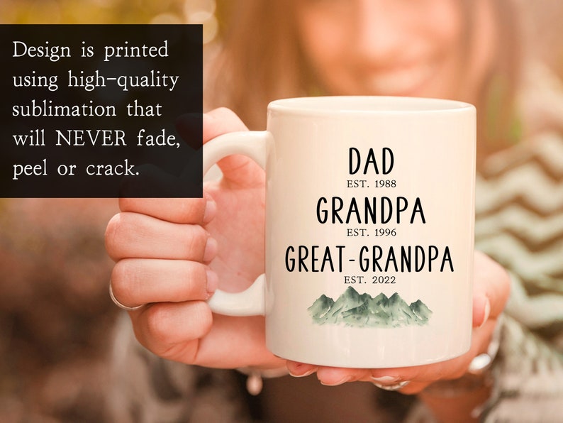 K&ouml;nnte beinhalten: Wei&szlig;e Keramiktasse mit schwarzem Text, der "DAD EST. 1988 GRANDPA EST. 1996 GREAT-GRANDPA EST. 2022" lautet, mit einem gr&uuml;nen Aquarell-Bergketten-Design am Boden.