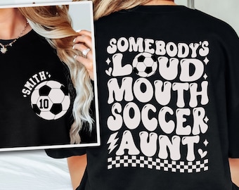 May include: Black t-shirt with white text that reads "Somebody's Loud Mouth Soccer Aunt" with a soccer ball graphic. The back of the shirt has a white soccer ball with the number 10 and the text "Smith" in a circle.