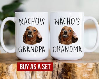 May include: Two white ceramic mugs with black text and images of a dachshund. The mug on the left says "Nacho's Grandpa" and the mug on the right says "Nacho's Grandma".
