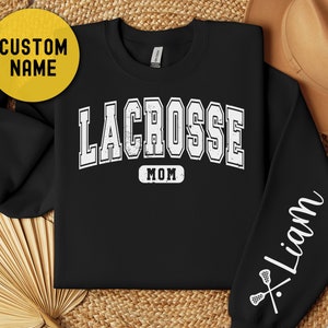 Op de afbeelding: Zwart sweatshirt met witte tekst die "Lacrosse Mom" zegt en een lacrosse stick en bal afbeelding op de mouw. De tekst "Liam" staat op de mouw.