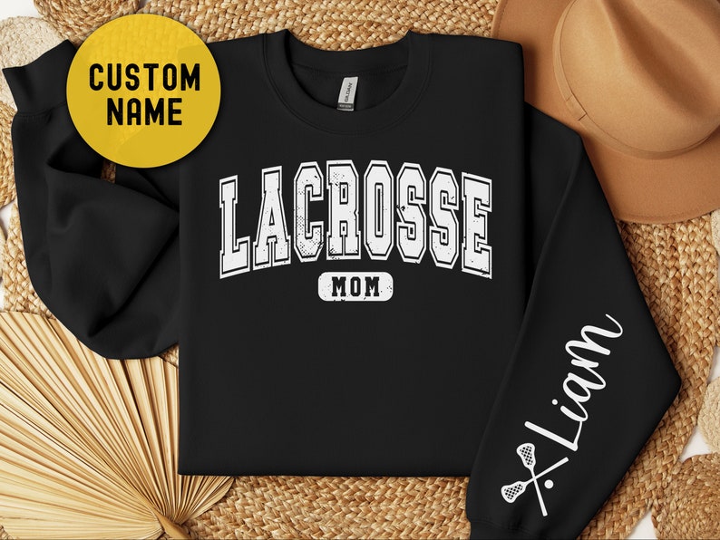 Op de afbeelding: Zwart sweatshirt met witte tekst die "Lacrosse Mom" zegt en een lacrosse stick en bal afbeelding op de mouw. De tekst "Liam" staat op de mouw.