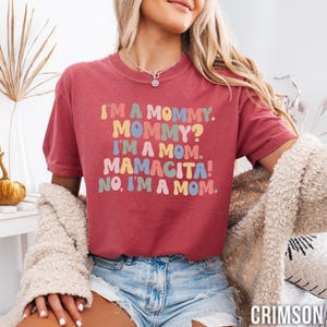 K&ouml;nnte beinhalten: Karmesinrotes T-Shirt mit dem Text "I'M A MOMMY. MOMMY? I'M A MOM. MAMACITA! NO, I'M A MOM." in farbenfrohen Buchstaben im Retro-Stil. Das Shirt hat einen Rundhalsausschnitt und kurze &Auml;rmel.