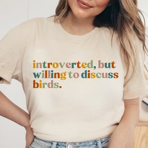 Op de afbeelding: Een wit T-shirt met de tekst "introvert, maar bereid om over vogels te praten." in kleurrijke letters.