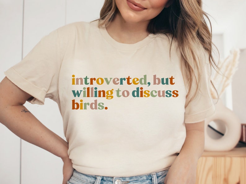 Op de afbeelding: Een wit T-shirt met de tekst "introvert, maar bereid om over vogels te praten." in kleurrijke letters.