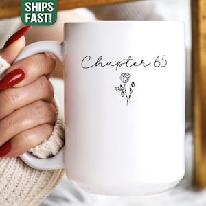 May include: White ceramic mug with the text "Chapter 65" in a handwritten font, accompanied by a small floral illustration. The mug is held by a person wearing a cream-colored sweater. The mug is a gift for a birthday.