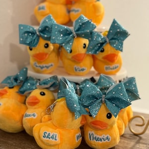 May include: A collection of yellow plush duck toys with teal glitter bows. Each duck has a name tag. Some ducks have a zipper on the back. The ducks are arranged on a tiered display.