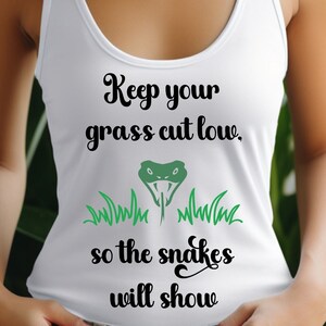 May include: White tank top with the text "Keep your grass cut low, so the snakes will show." The design includes a green snake head and green grass illustrations. The text and graphics are in black and green.