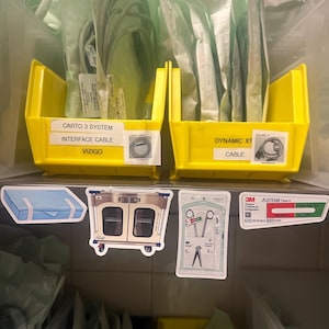 May include: Two yellow plastic storage bins containing medical cables in sterile packaging. Below, four stickers are displayed: a blue box, a medical cart, a pair of surgical scissors in a pouch, and a 3M Steam Chemical Integrator label.