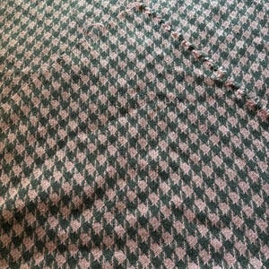 May include: Close-up of a green and tan houndstooth fabric. The fabric has a repeating pattern of small, diamond-shaped designs. The edges of the fabric are frayed in some areas.