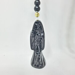 May include: A black figurine depicting a hooded skeletal figure with a skull face, draped in a long robe. The figure is suspended from a string featuring matte black spherical beads and a single polished gold-tone bead positioned above the head.