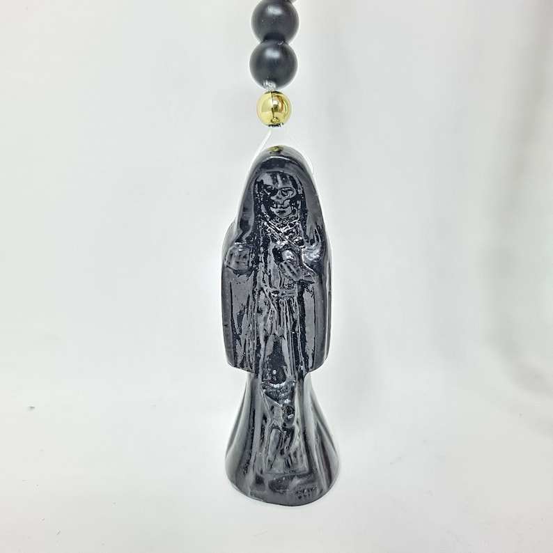 May include: A black figurine depicting a hooded skeletal figure with a skull face, draped in a long robe. The figure is suspended from a string featuring matte black spherical beads and a single polished gold-tone bead positioned above the head.