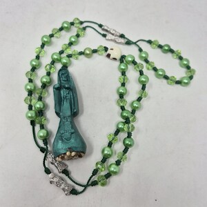 May include: A rosary featuring light green pearl-like beads and faceted lime green beads strung on dark green cord. The piece includes a dark teal figure pendant, two silver-toned metal charms, and a small off-white skull bead near the top.