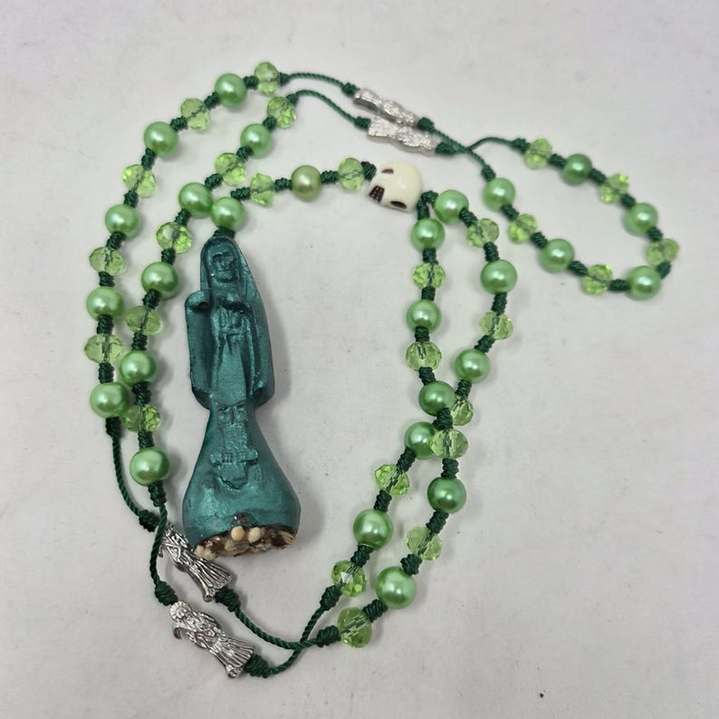 May include: A rosary featuring light green pearl-like beads and faceted lime green beads strung on dark green cord. The piece includes a dark teal figure pendant, two silver-toned metal charms, and a small off-white skull bead near the top.