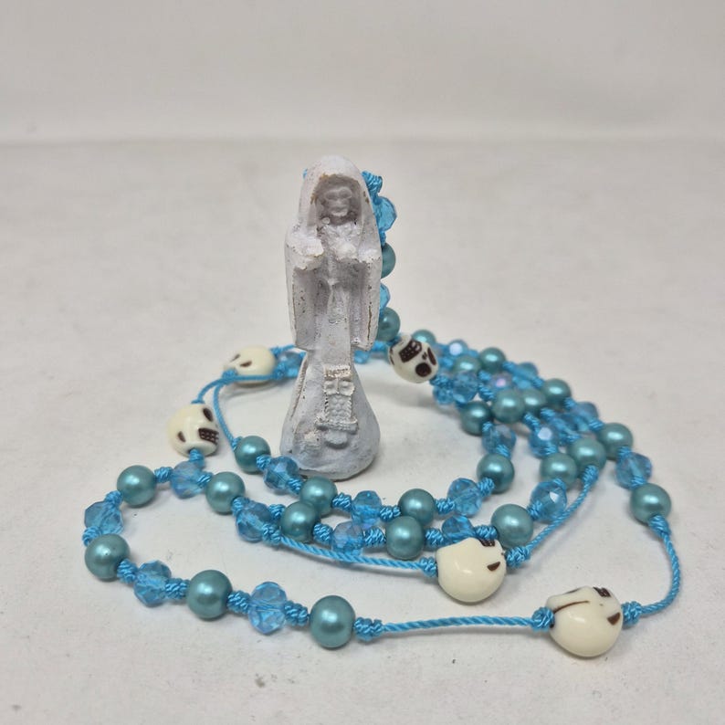 May include: A white figurine of a hooded figure standing upright, surrounded by a long string of light blue beads, translucent blue faceted crystals, and small off-white skull-shaped beads, all connected by a bright blue knotted cord.