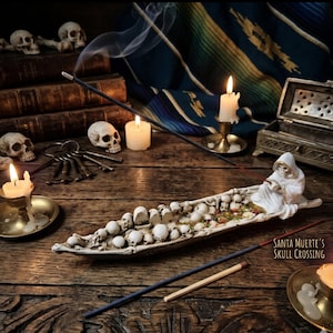 May include: An off-white resin incense burner shaped like a boat filled with skulls, featuring a hooded skeletal figure at the stern. The burner has the text SANTA MUERTE'S SKULL CROSSING printed on the side and holds a lit incense stick.