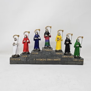 May include: Seven Santa Muerte figurines, each representing a different power: Protection, Love, Health, 7 Powers, Money, Against Damage, Legal Case. Each figurine is a skeletal figure wearing a robe in a different color: white, red, blue, rainbow, yellow, black, and green. The figurines are arranged on a tiered platform with the name of each power written below.