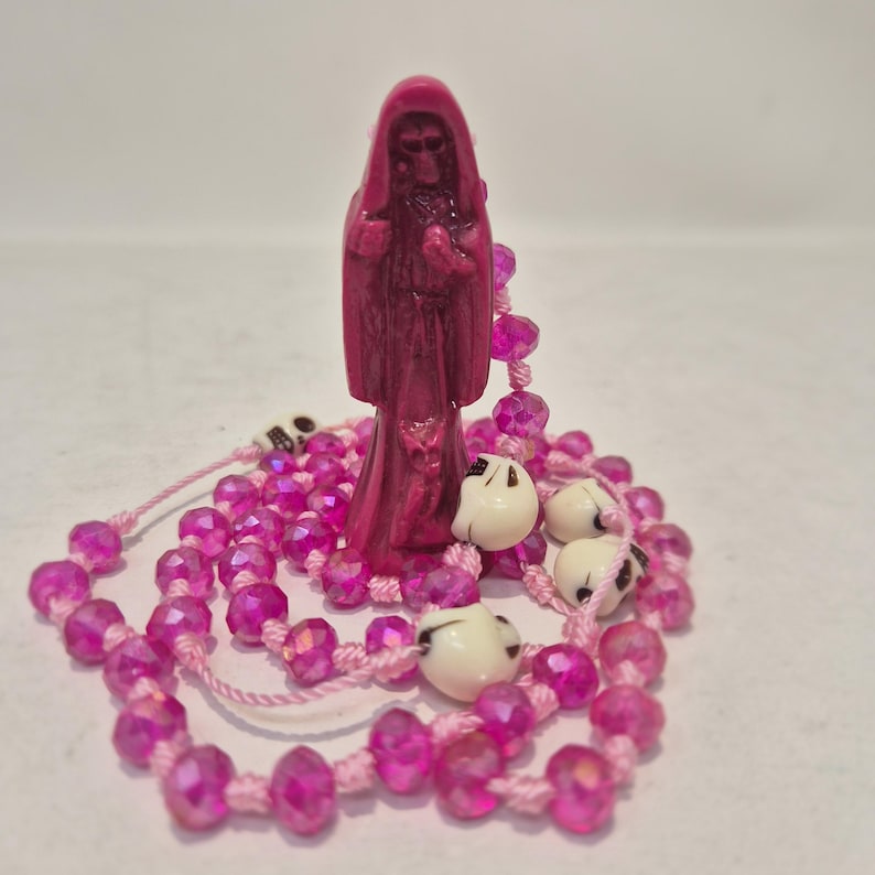 May include: A magenta-colored figurine of a hooded figure standing upright, surrounded by a rosary-style string of faceted pink beads and several small, off-white skull-shaped beads interspersed throughout the circular arrangement of the cord.