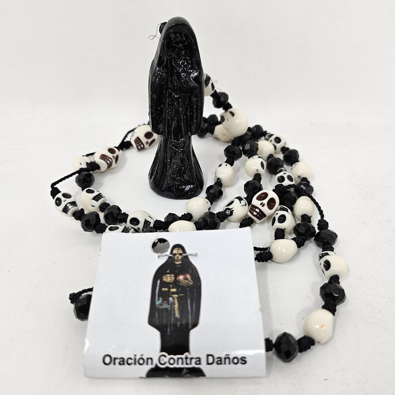 May include: A black figurine of a hooded figure standing behind a rosary made of white skull beads and black faceted beads. A white rectangular card is attached, featuring an illustration of the figure and the text Oraci&oacute;n Contra Da&ntilde;os.