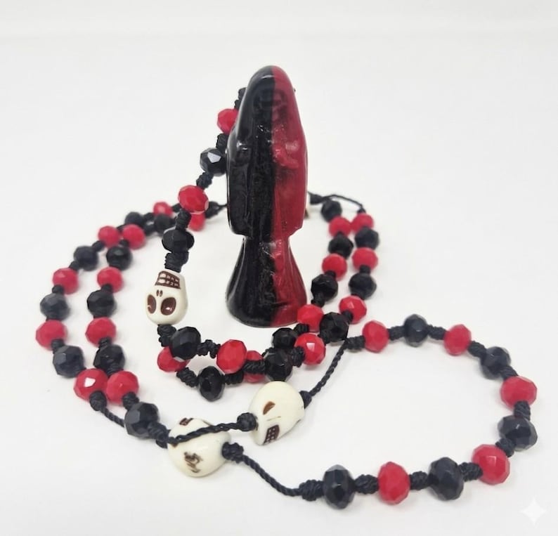 May include: A black and red figurine stands upright, surrounded by a beaded necklace featuring alternating black and red faceted beads. The necklace includes three off-white skull-shaped beads integrated into the black cord stringing.